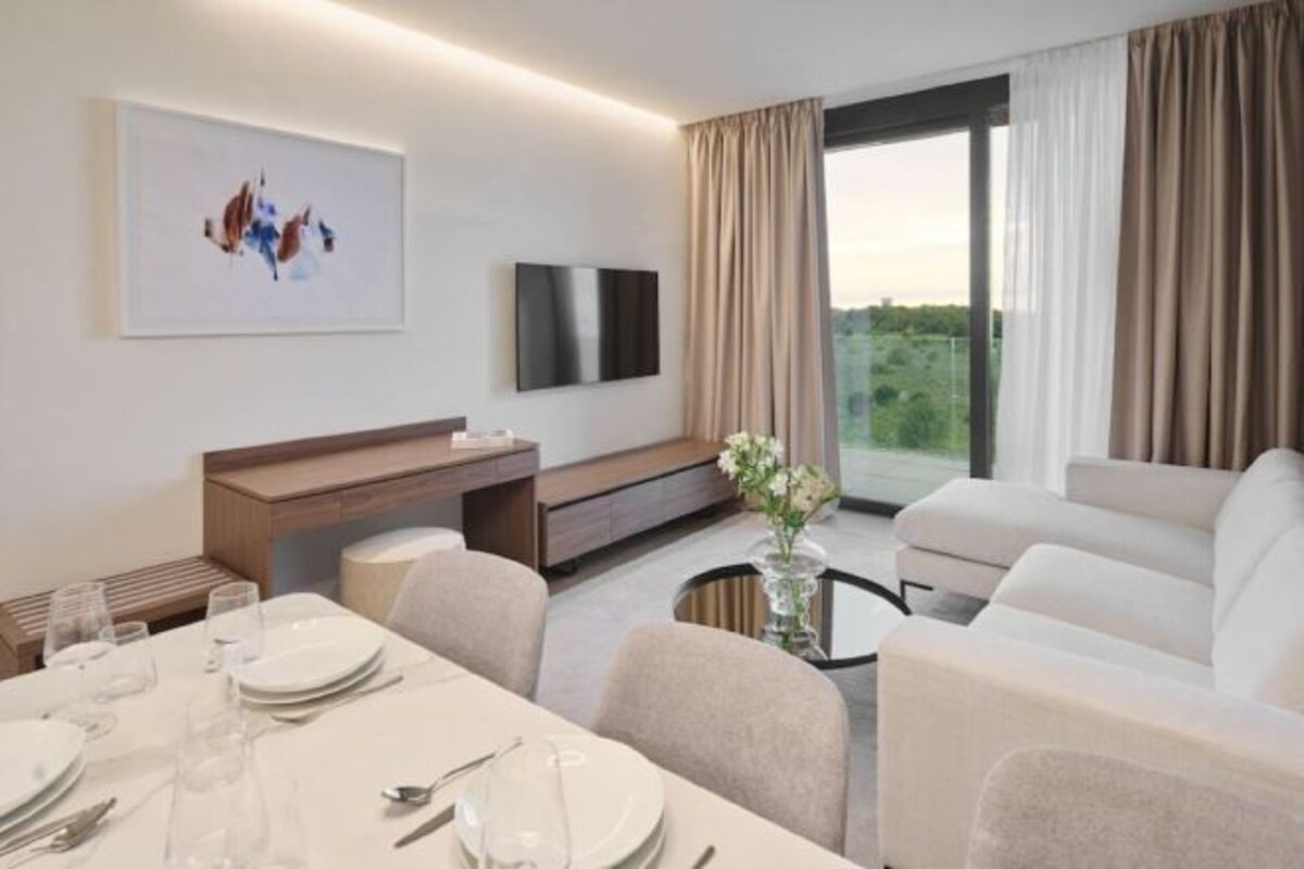 Habitación de hotel moderna con zona de comedor, televisor de pantalla plana, sofá y un amplio ventanal con cortinas.