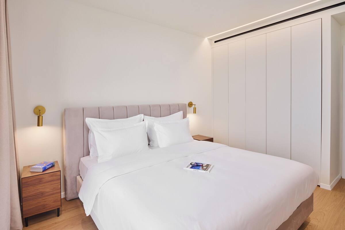 A neatly made bed with white linens, a wooden nightstand with a book, and a wall-mounted light fixture.