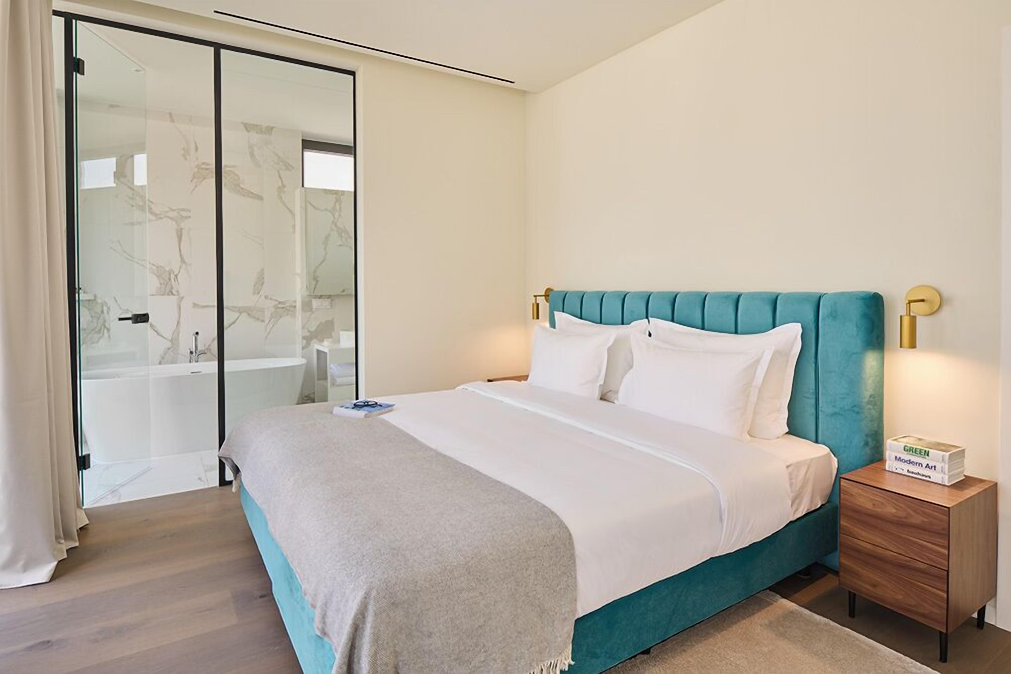 A modern bedroom with a teal upholstered headboard, a white bedspread, and a wooden nightstand with a box labeled "GREEN Modern Art".
