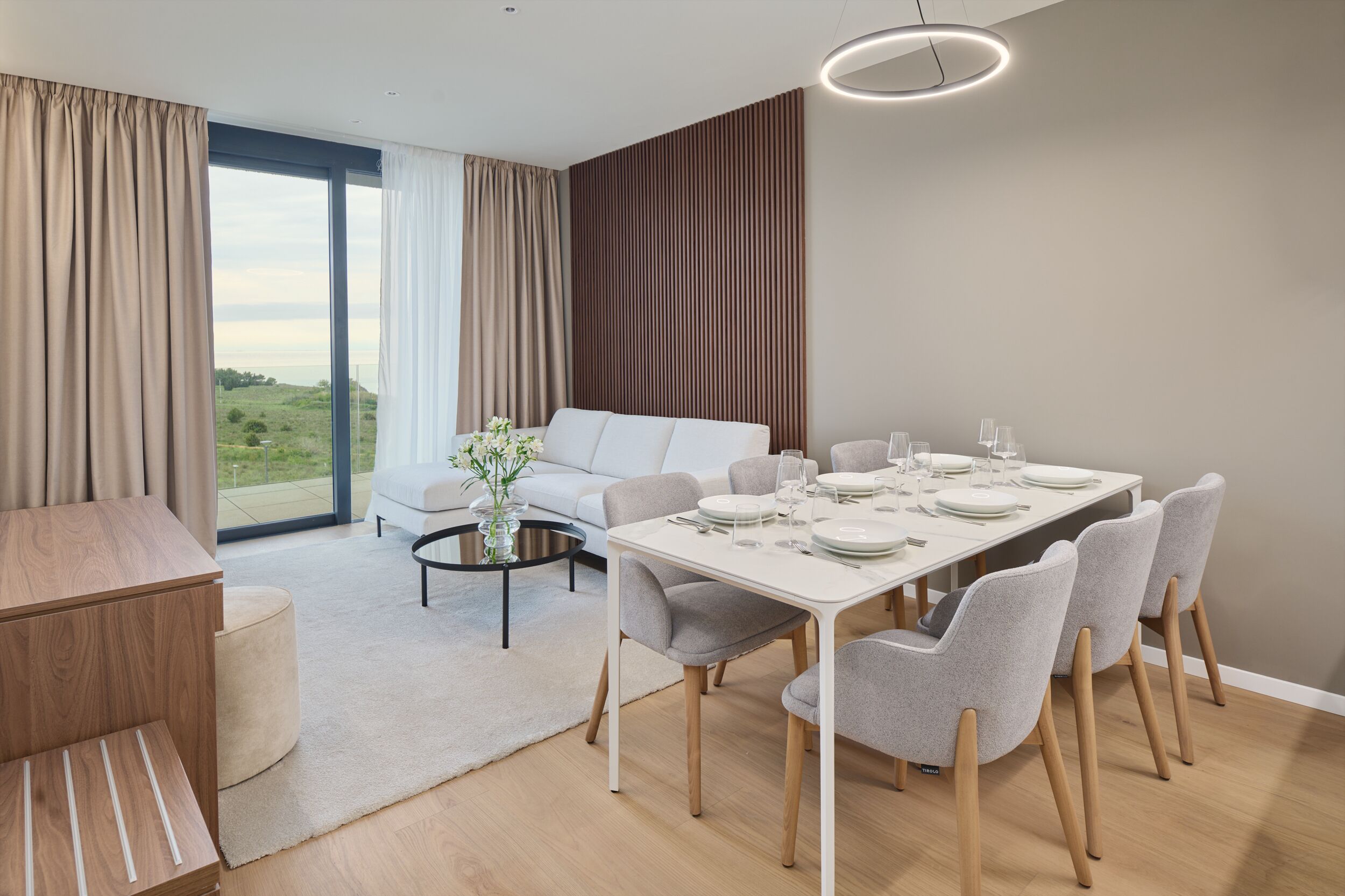 A modern dining area with a white table set for six, surrounded by grey chairs. A white sofa and a round coffee table with a vase of flowers are in the background.