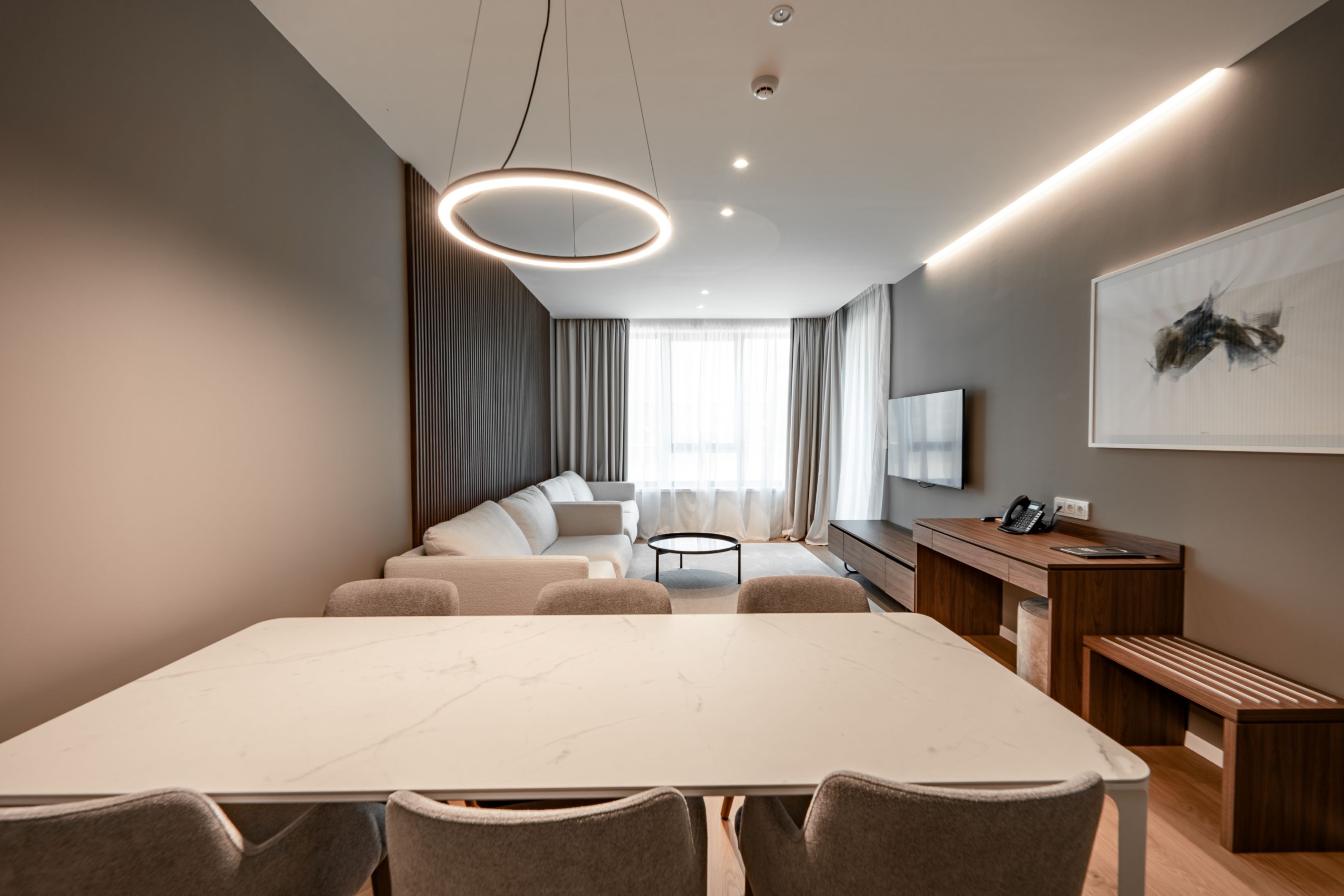 A modern hotel room with a large marble dining table, a sofa set, a wooden desk, and a large abstract painting on the wall.