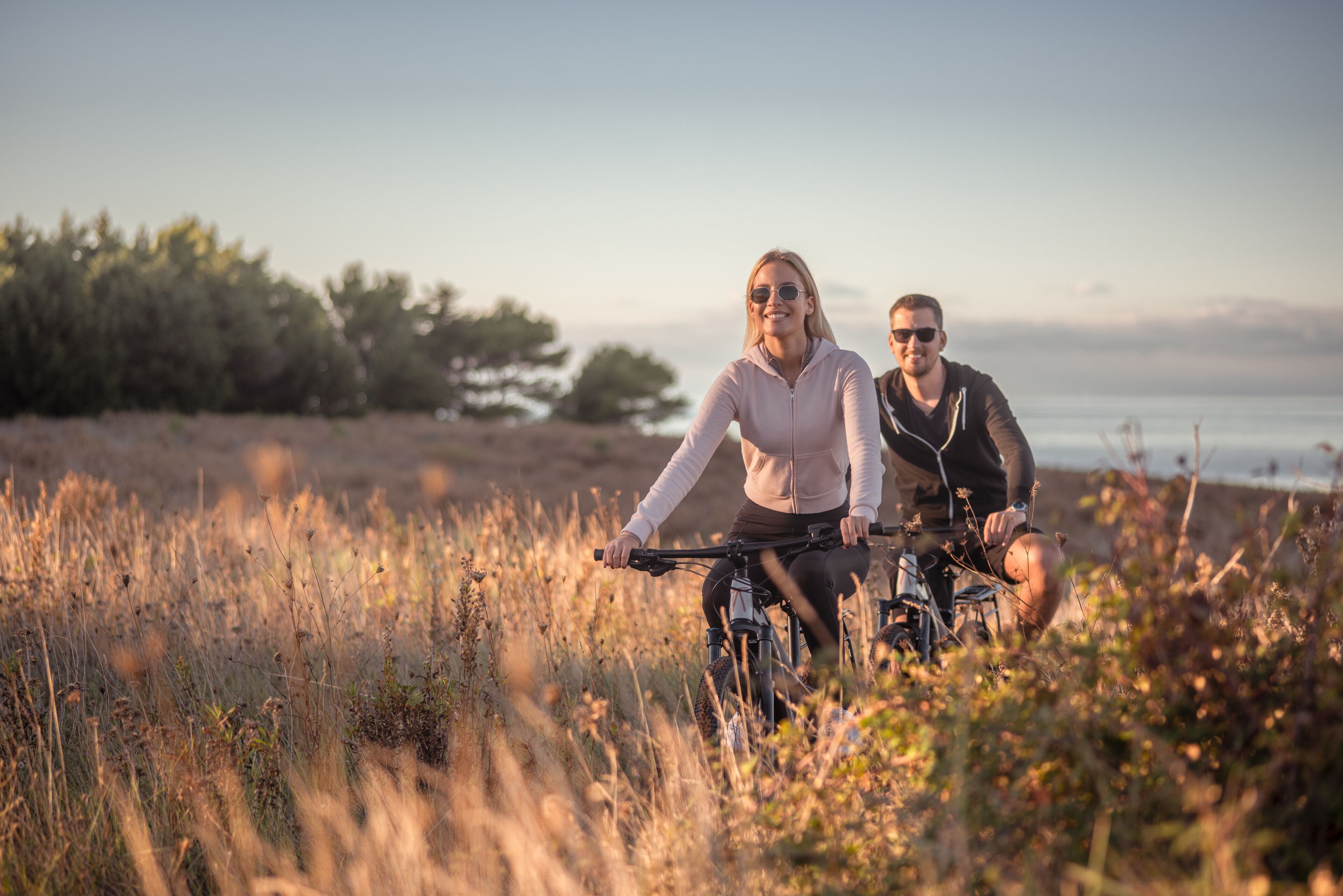 Dos personas en bicicleta en un entorno natural, con árboles y una masa de agua al fondo.