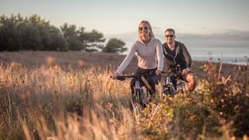 Dos personas en bicicleta en un entorno natural, con árboles y una masa de agua al fondo.