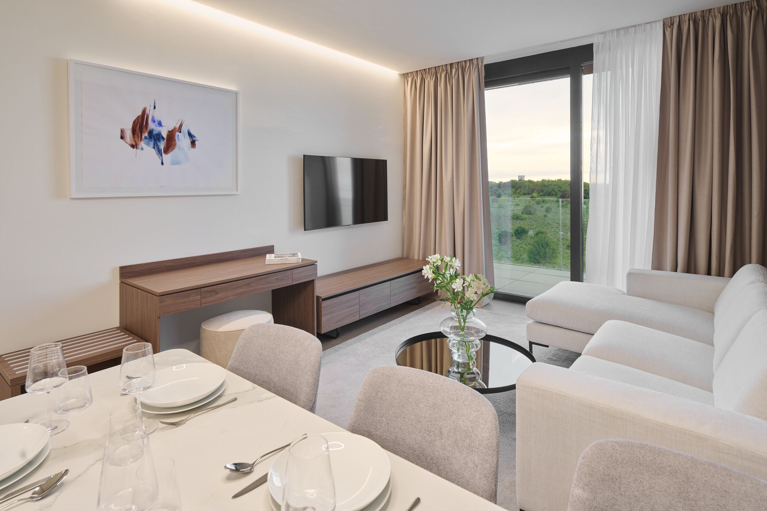 Una habitación de hotel moderna con área de comedor, un sofá, un televisor y un ventanal con cortinas.