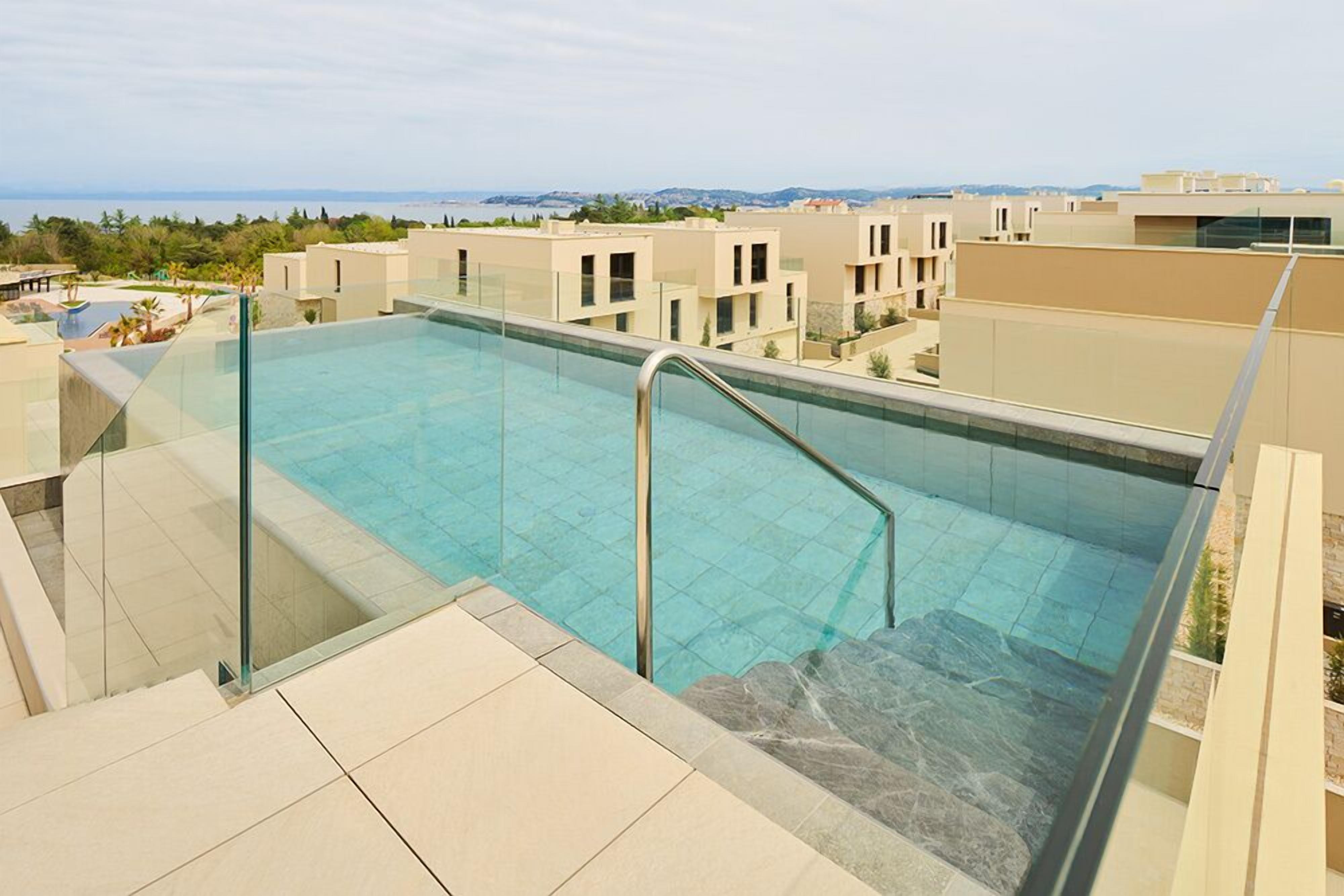 A rooftop infinity pool with clear blue water, surrounded by beige buildings and a view of the sea.