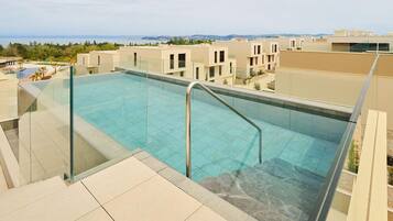 A rooftop infinity pool with clear blue water, surrounded by beige buildings and a view of the sea.