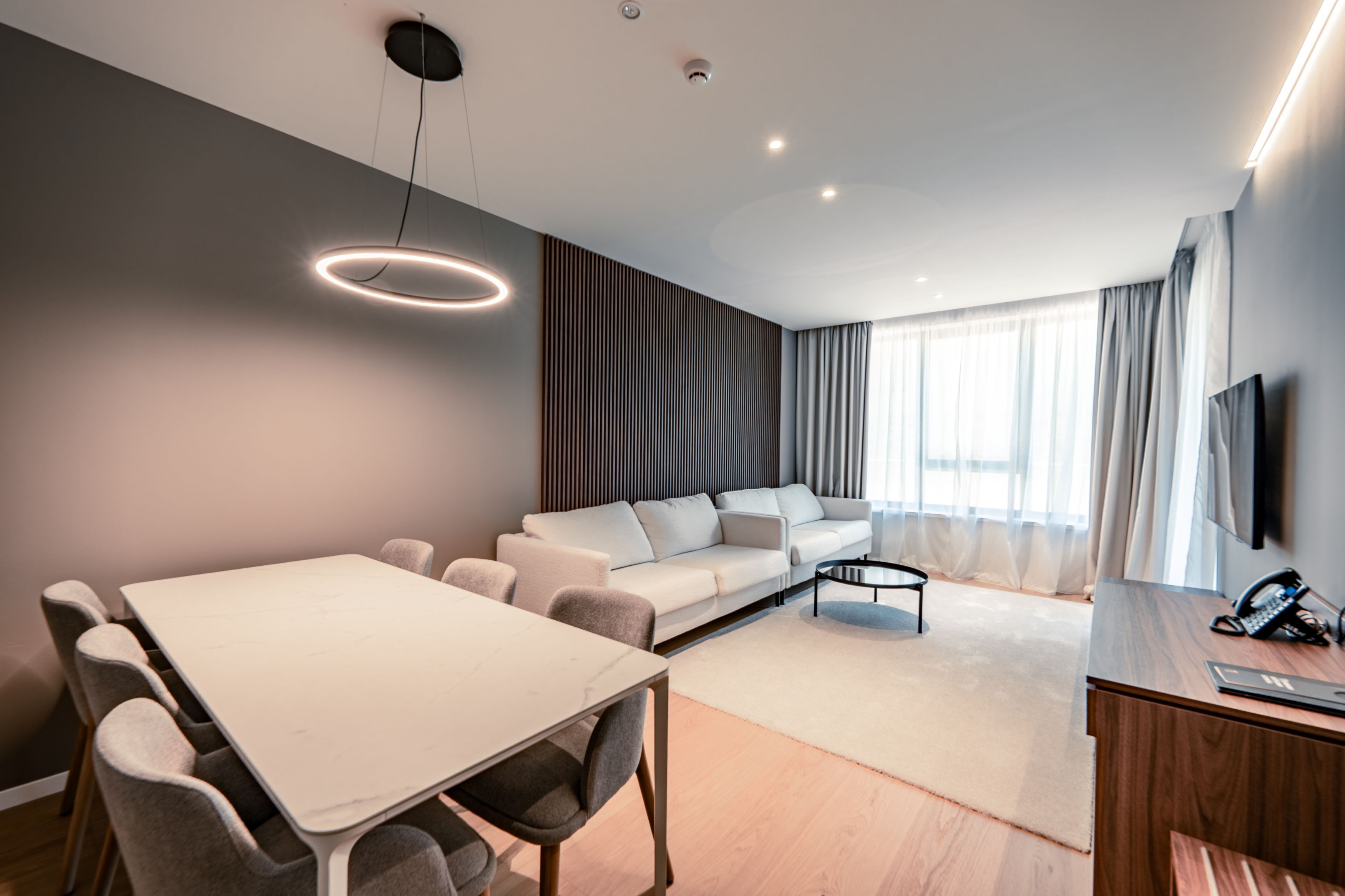 A modern living room with a white sofa, a dining table with chairs, a coffee table, and a TV mounted on the wall.