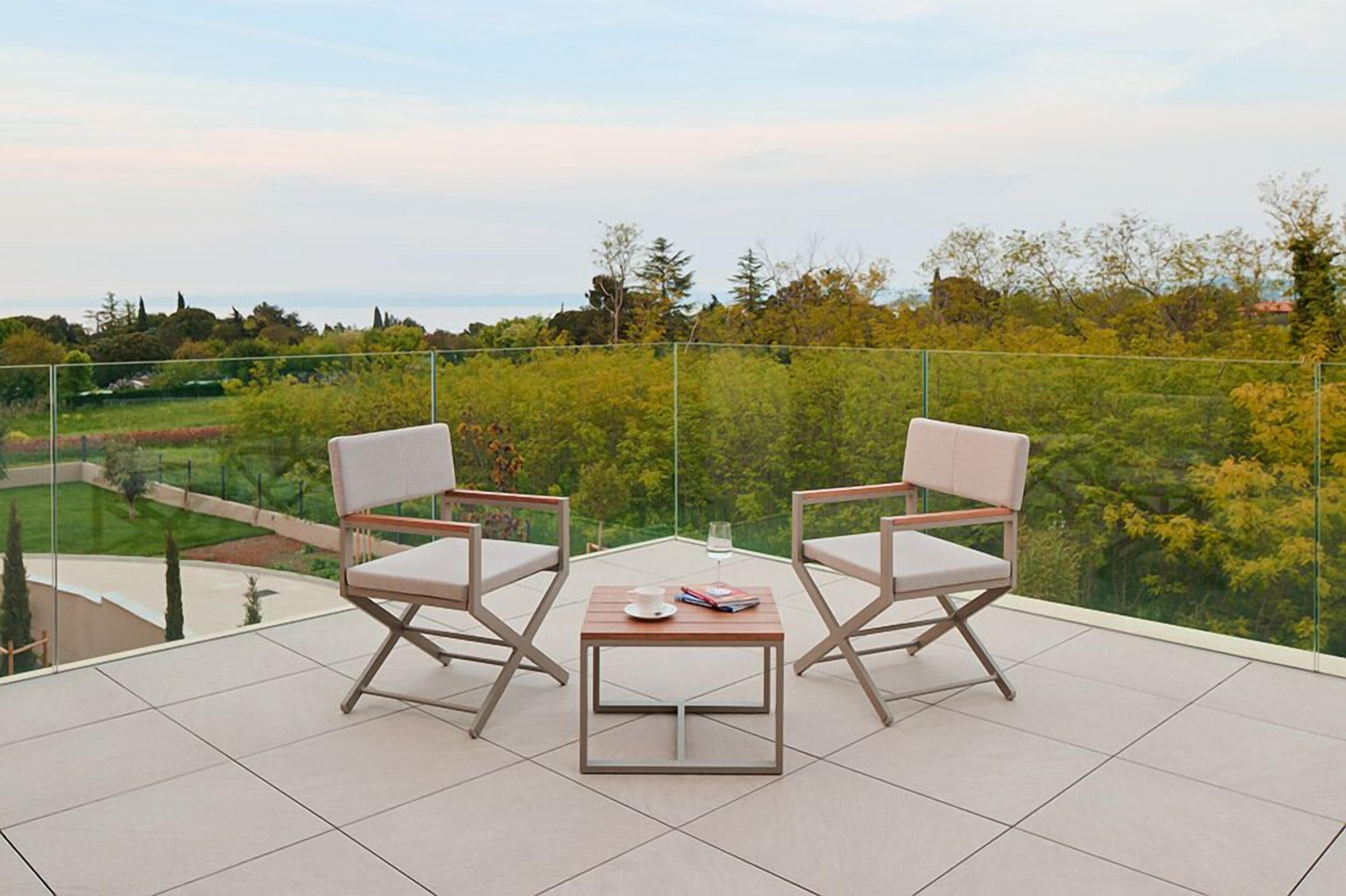 Varanda com duas cadeiras e uma mesa, com vista para um jardim exuberante e árvores ao longe.