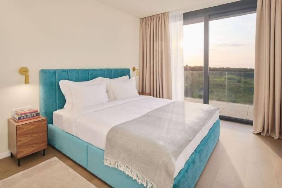 A bedroom with a large bed, a nightstand with books, and a view of the outdoors.