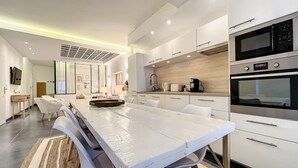 Een moderne keuken met witte kastjes, roestvrijstalen apparatuur en een lange eettafel met stoelen.