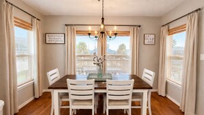 Une salle à manger avec une table en bois, des chaises blanches, un lustre et deux fenêtres avec des rideaux.