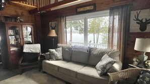 Ein gemütliches Wohnzimmer mit einer Couch, einem Sessel, einem Bücherregal und einem Fenster mit Blick auf Bäume.