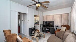 Una sala de estar con ventilador de techo, televisor, sofá, mesa de centro y una cocina al fondo.