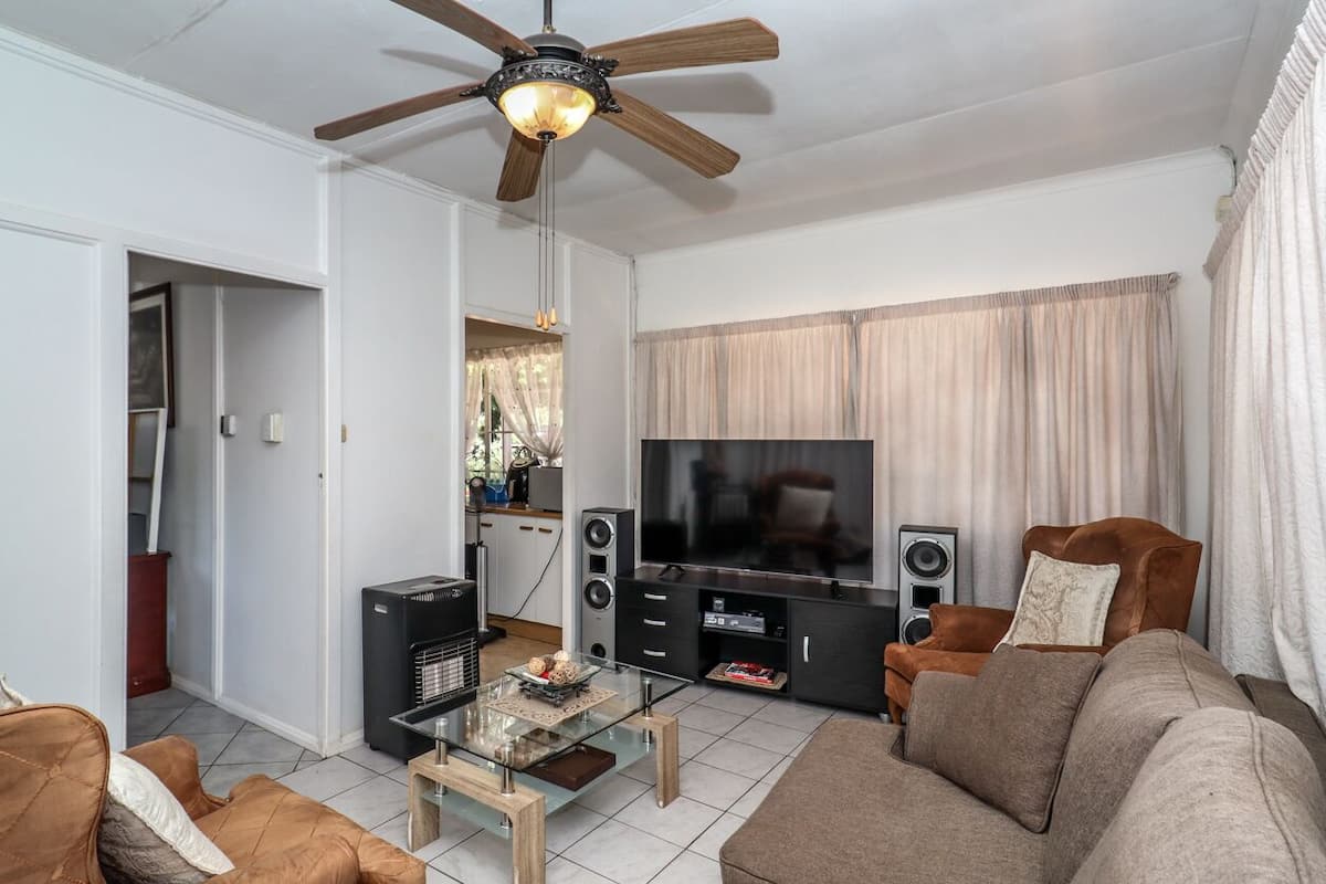 A living room with a ceiling fan, a TV, a sofa, a coffee table, and a kitchen area in the background.