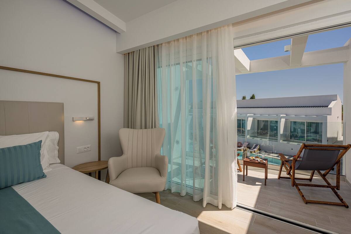 Habitación de hotel con cama, una silla, una mesita, balcón con piscina y vistas del edificio.
