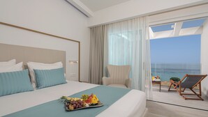 Een hotelkamer met een bed, een schaal fruit, een stoel en een balkon met uitzicht op zee.