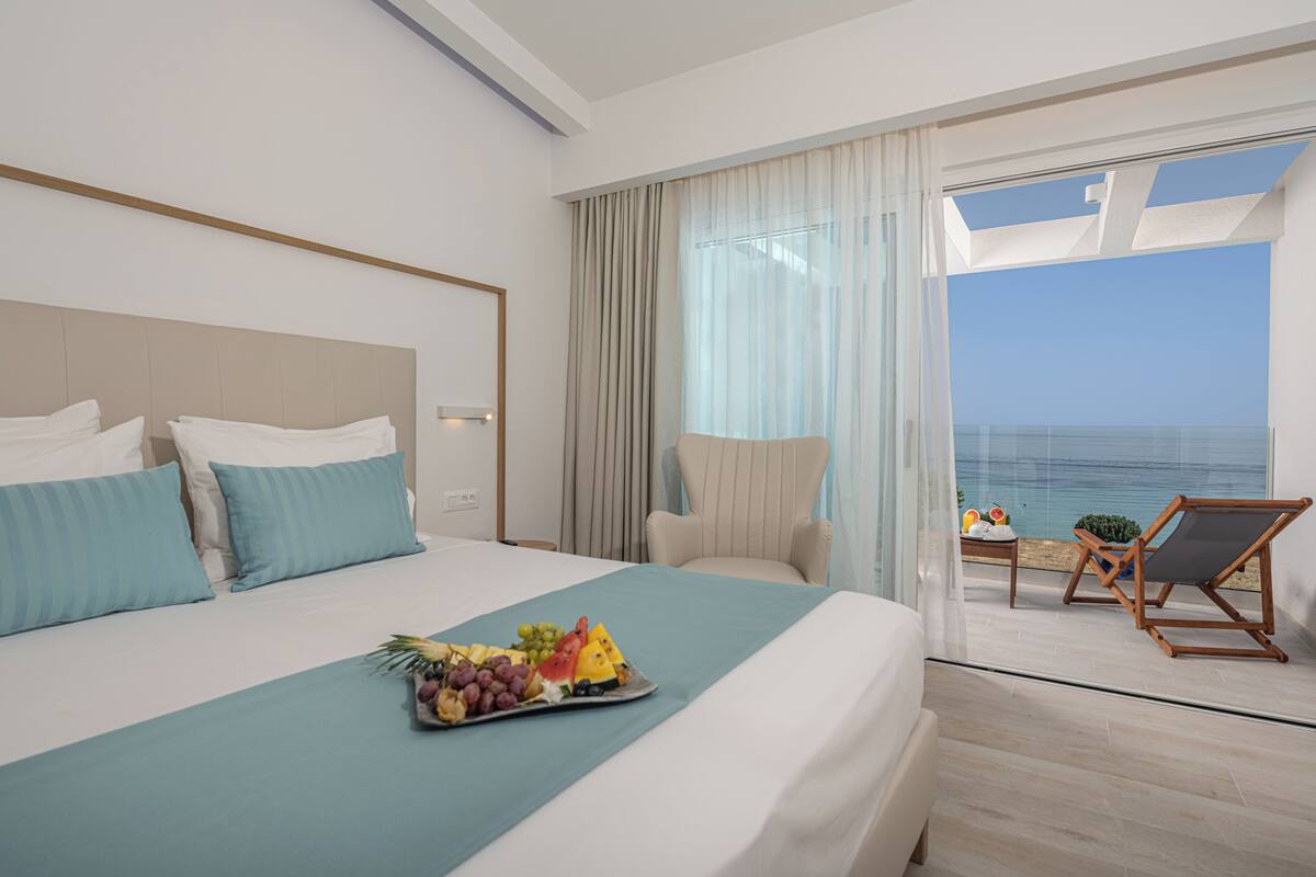 Habitación de hotel con cama, bandeja de frutas, una silla y balcón con vistas al mar.