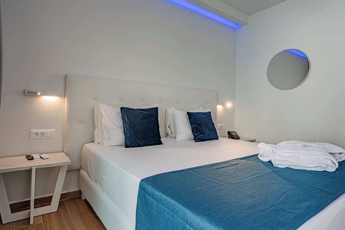 Una habitación de hotel moderna con una cama bien hecha, una mesita de noche con teléfono y control remoto, y una pequeña mesa con lámpara.