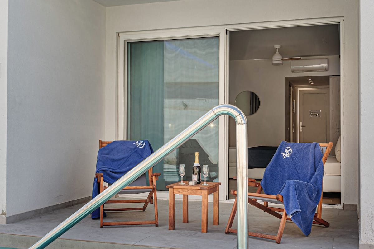 A pool area with a glass barrier, two covered chairs, a table with a bottle and glass, and a door leading to a room.