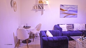 Een moderne woonkamer met een paarse bank, witte stoelen, een glazen salontafel en een wandplank met boeken en decoratieve voorwerpen.