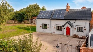 A white cottage with a red door, solar panels on the roof, and a fenced yard. - Tumbleweed Cottage (Newark)