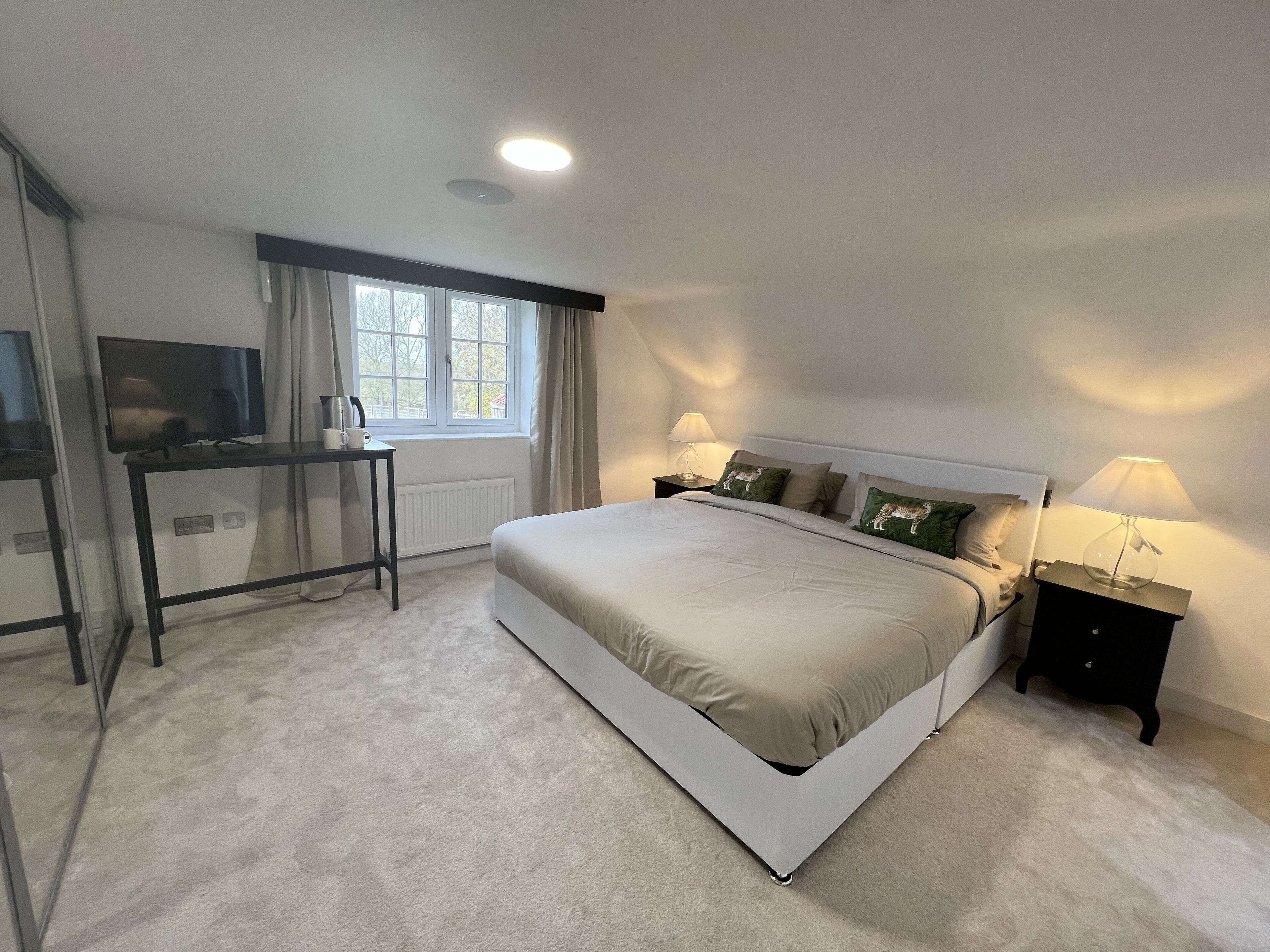 Quarto com cama, mesas de cabeceira, escrivaninha, televisão e janela com cortinas.