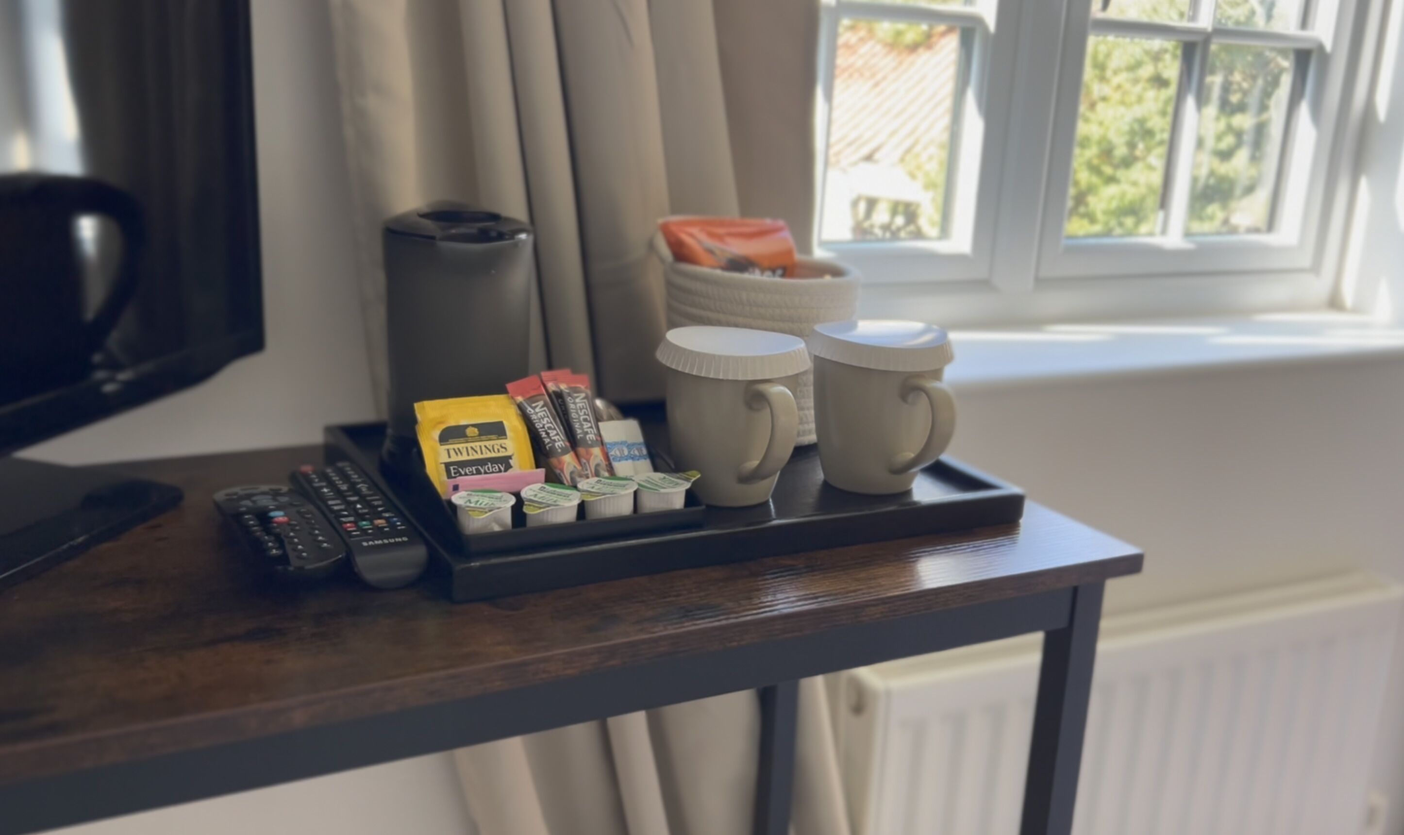 A tray with coffee mugs, a remote control, and coffee pods on a wooden table near a window.