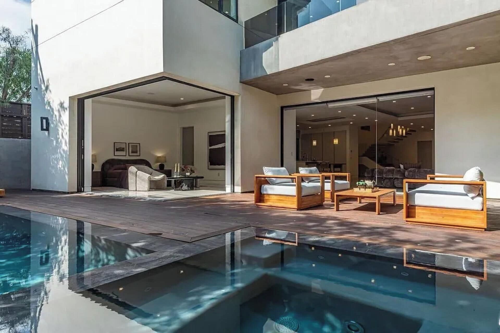 Un área de piscina moderna al aire libre con un deck de madera, sillas de descanso y un espacio habitable con paredes de vidrio.