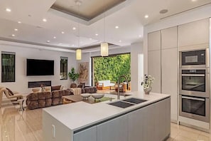A modern kitchen with a central island, built-in ovens, and a dining area with a view of a green wall.