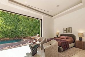 A bedroom with a large window, a bed, a nightstand, a chair, and a small table with a vase.