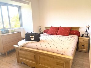 Une chambre avec un lit en bois, des coussins à pois rouges, une table de chevet avec une lampe et une fenêtre donnant sur un paysage verdoyant.