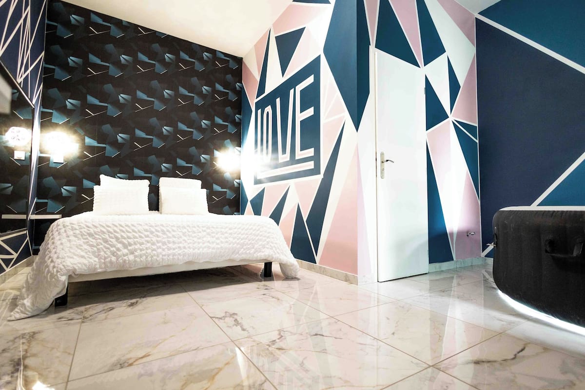 A modern bedroom with a geometric-patterned wall, a bed with white bedding, and a large mirror.