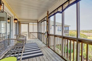 A covered porch with wicker chairs, a striped rug, and a view of a house and landscape. - Marsh Mellow | Soundfront+Private Dock+Beach Access (Surf City)