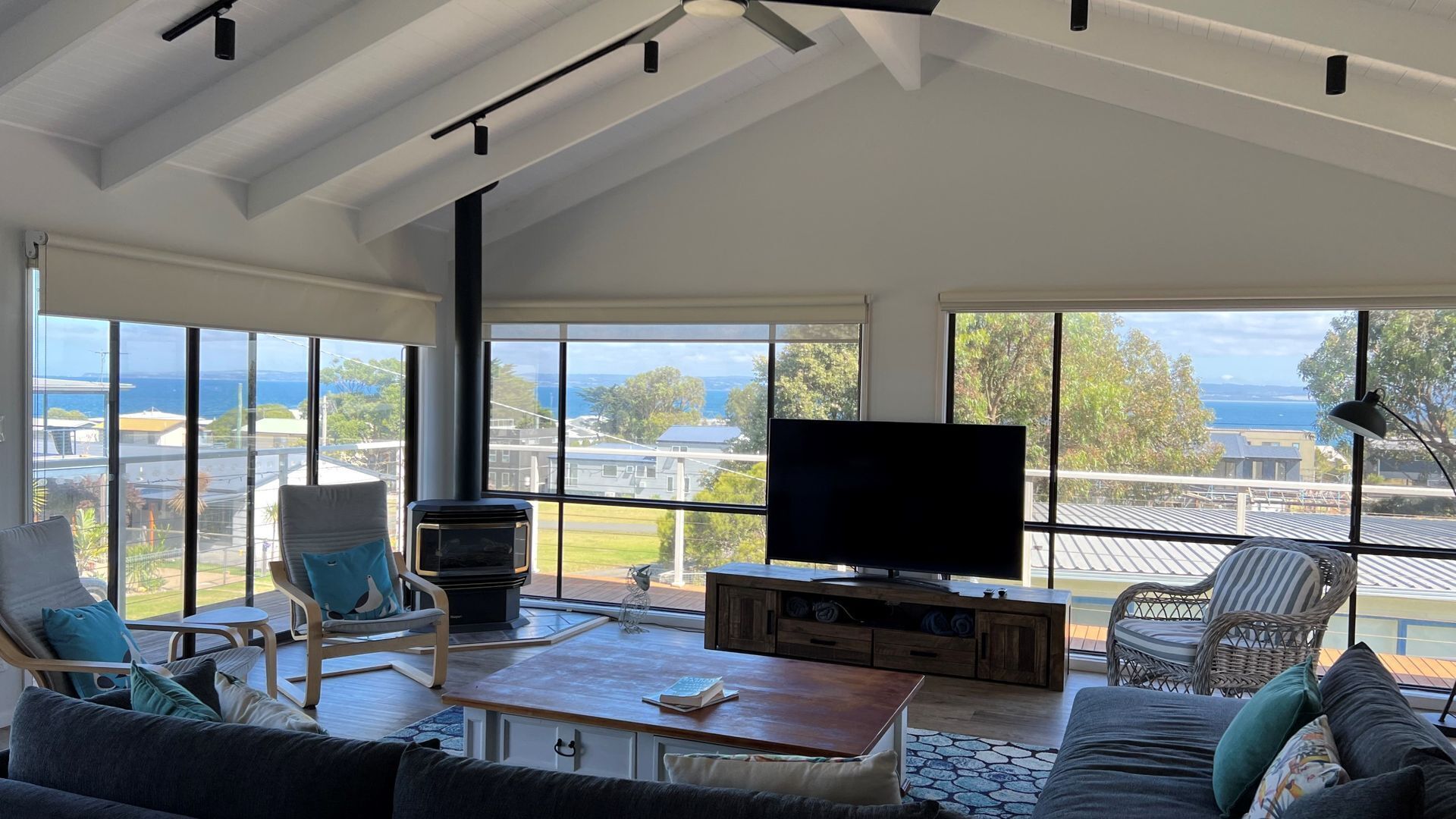 A modern living room with a large TV, comfortable seating, and a view of the sea.