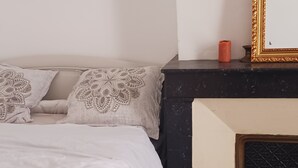 A bed with a white duvet and decorative pillows, a black marble fireplace, and a framed picture leaning against the wall. - Appartement Centre Ville 110m2 (Béziers)