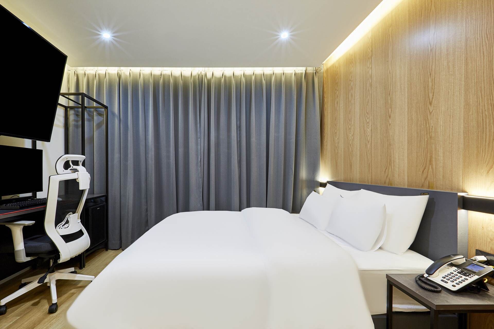 A hotel room with a bed, bedside table, and a desk with a computer and chair.