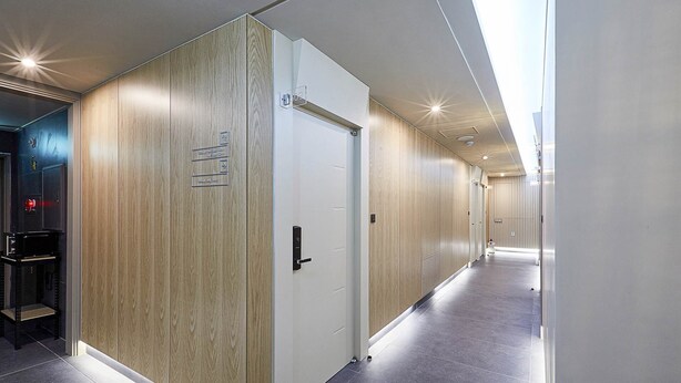 Un pasillo moderno de hotel con paneles de madera en las paredes, una puerta blanca con manija e indicaciones de números de habitación.
