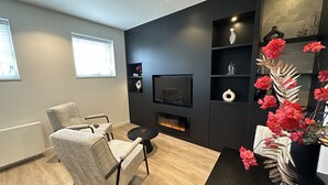 Un salon moderne avec une cheminée, un téléviseur fixé au mur et un coin salon comprenant un fauteuil et une table.