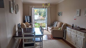 Una sala de estar con sofá, una mesa de comedor con sillas, una zona de cocina con armarios y un ventanal con cortinas.