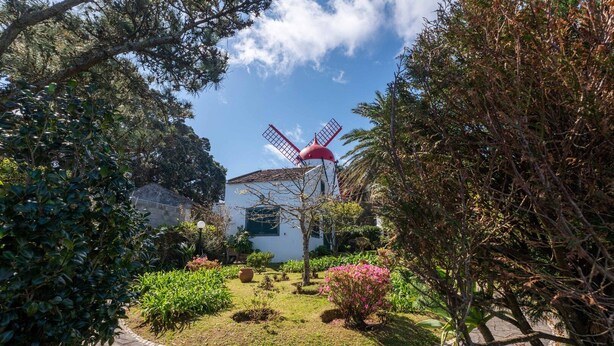 Un molino con techo rojo, rodeado de vegetación exuberante y un edificio blanco con tejado de tejas.