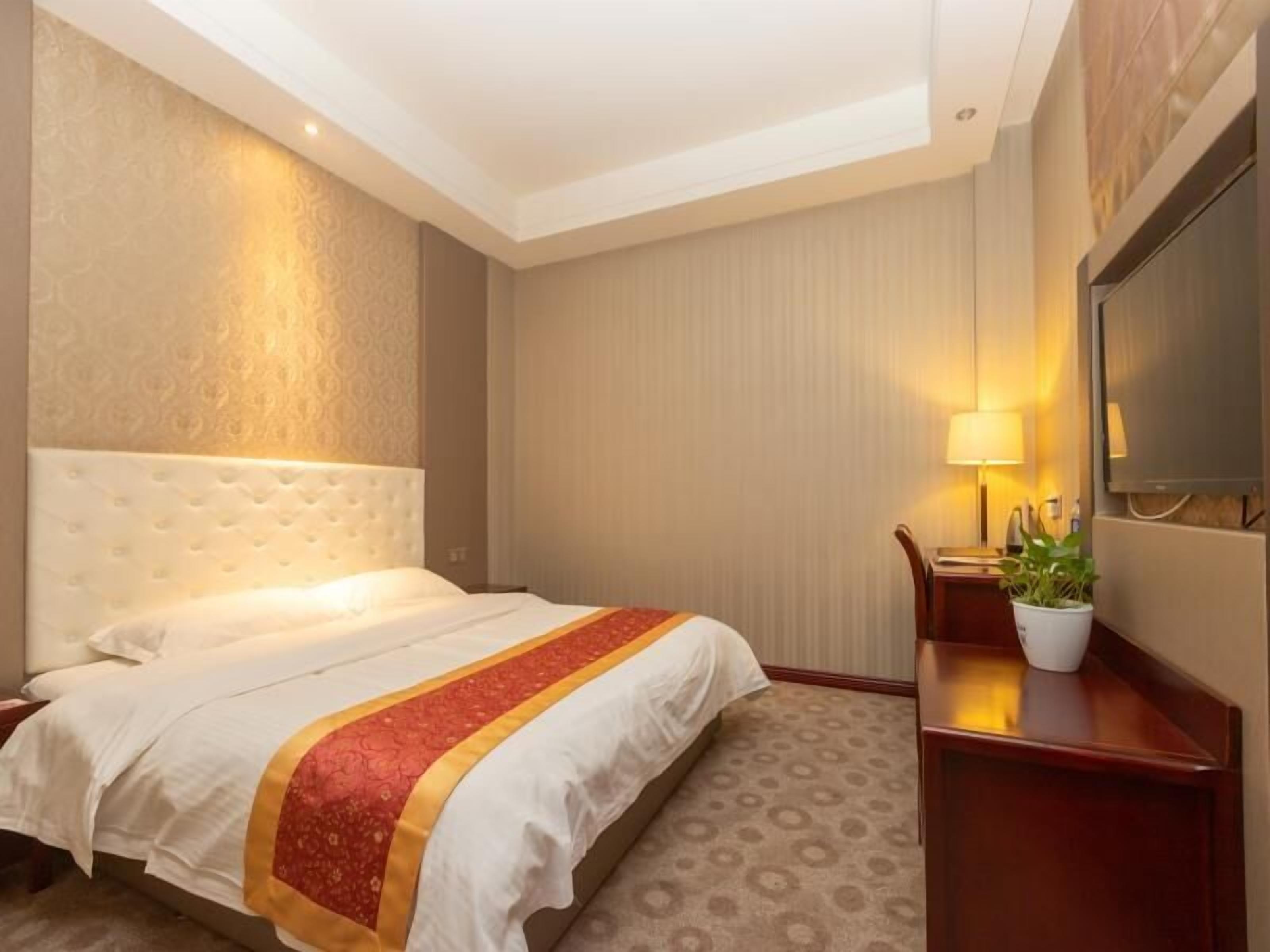 A hotel room with a large bed, a desk with a lamp, and a television on the wall.