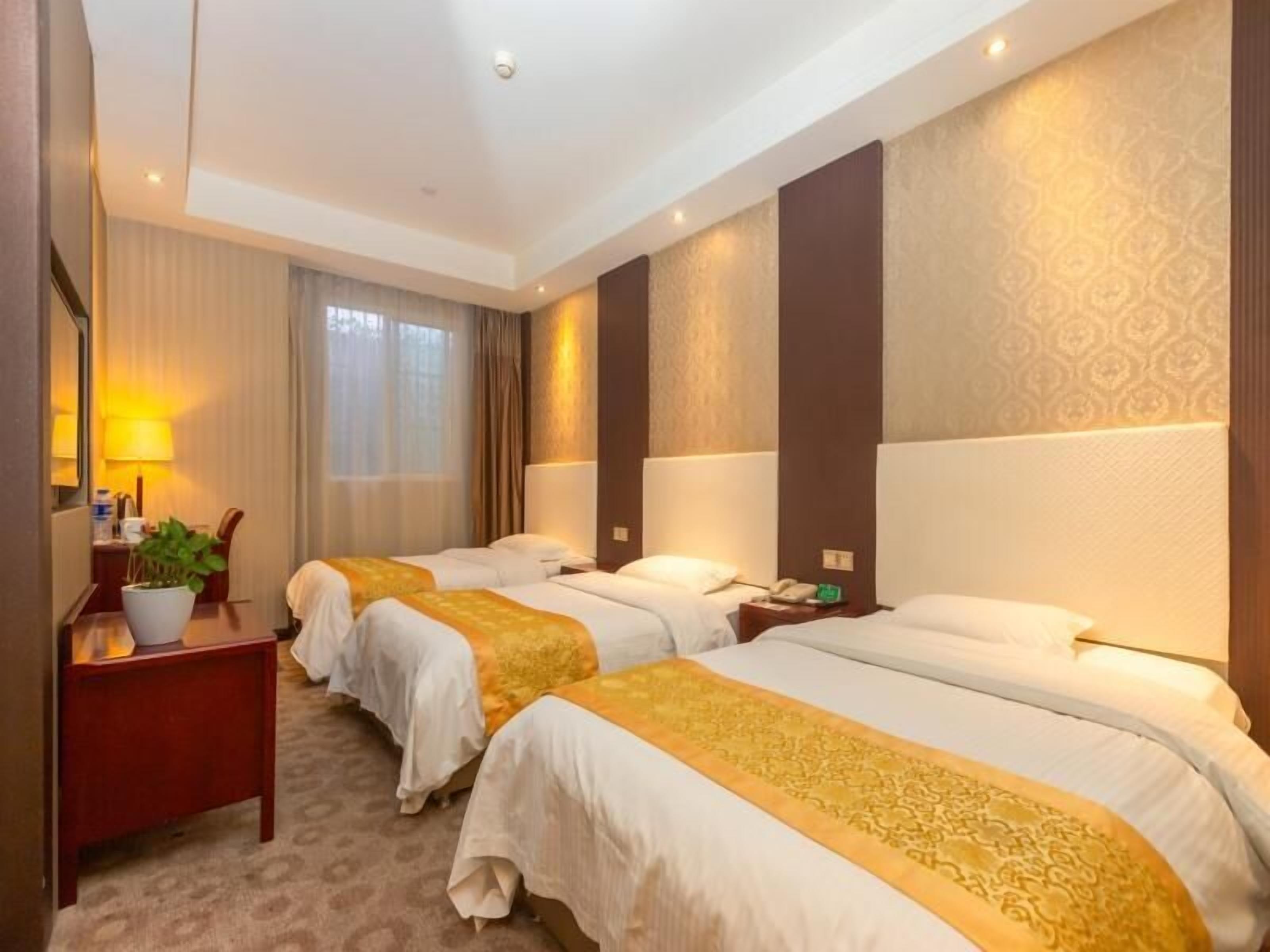 A hotel room with three beds, a desk with a lamp, and a potted plant.