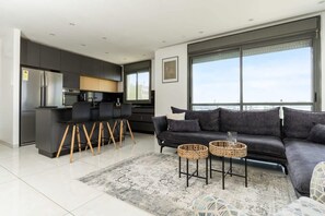 A modern living room with a grey sofa, two woven side tables, and a large window offering a view of the city. - Luxury  3 Bedrooms apartment in Baka (Jerusalem)