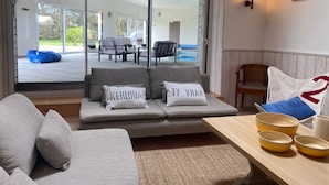 Un soggiorno moderno con un divano, cuscini e un tavolino. Una porta scorrevole in vetro dà accesso a una zona relax esterna con sedie e una piscina.