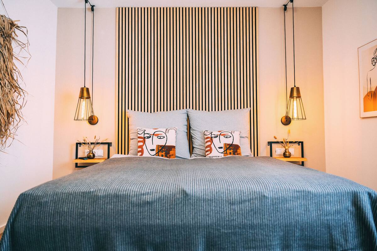 Uma cama bem arrumada com cabeceira listrada, duas mesas de cabeceira com abajures e uma prateleira na parede com objetos decorativos.