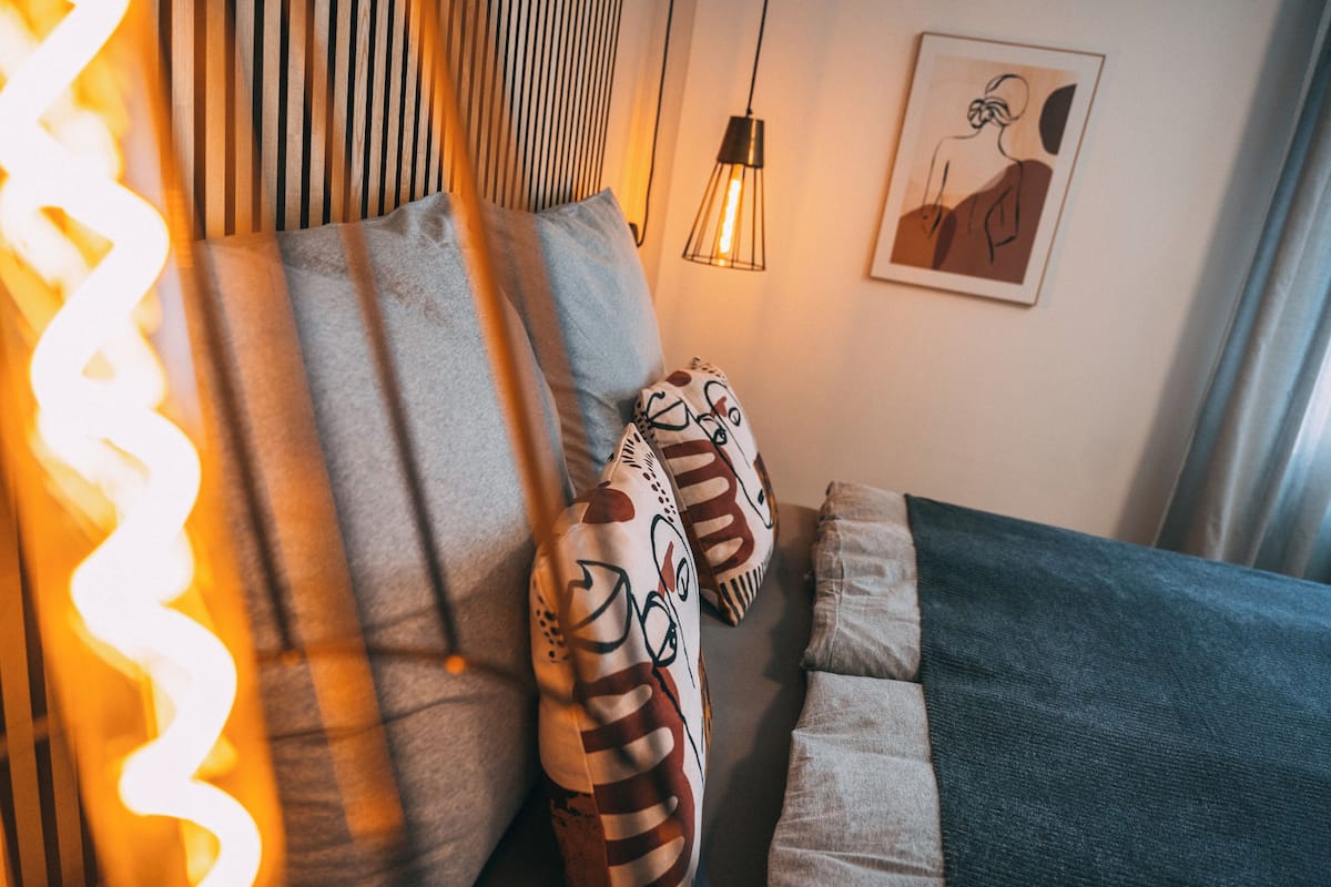 Um quarto acolhedor com cama, almofadas, luminária pendente e uma obra de arte emoldurada na parede.