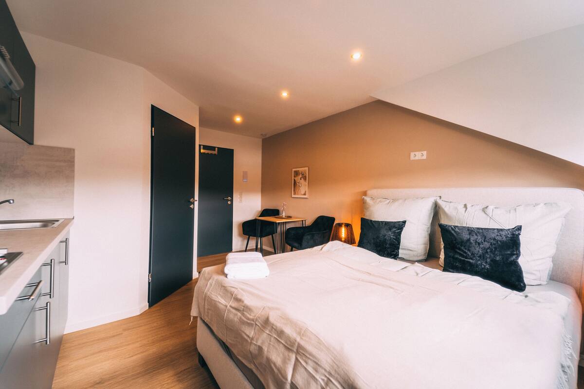 Quarto de hotel moderno com cozinha compacta, cama com almofadas brancas e pretas, e escrivaninha com cadeira.