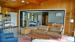 A modern living room with a large leather sofa, a blue armchair, and a wooden coffee table. There's a kitchen area with a white fridge and a dining table.