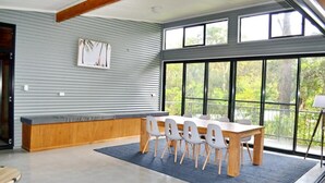 A modern dining area with a wooden table and white chairs, large windows with a view of greenery, and a bench along the wall.