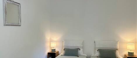 A bedroom with two single beds, each with a nightstand and a lamp. A mirror hangs on the wall to the left.